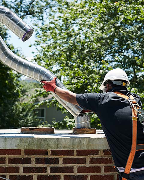 Technician connecting new stainless steel chimney lining to chimney.  He is wearing safety gear with trees in the background.