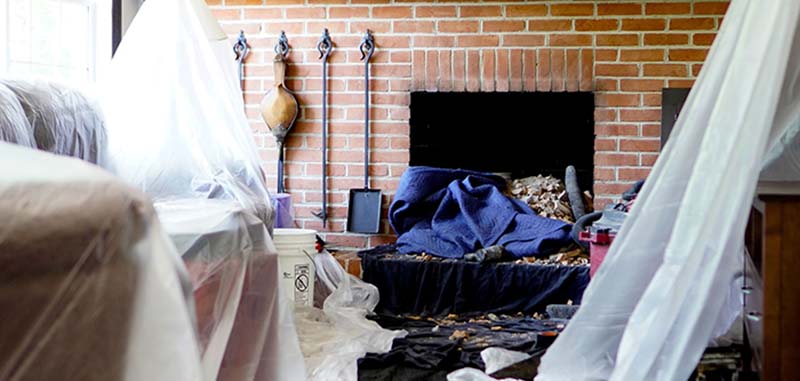 Winstons Chimney Original Photo of Fireplace Damage Damaged fireplace with blue blanket draped over rubble on hearth and plastic sheeting covering furniture