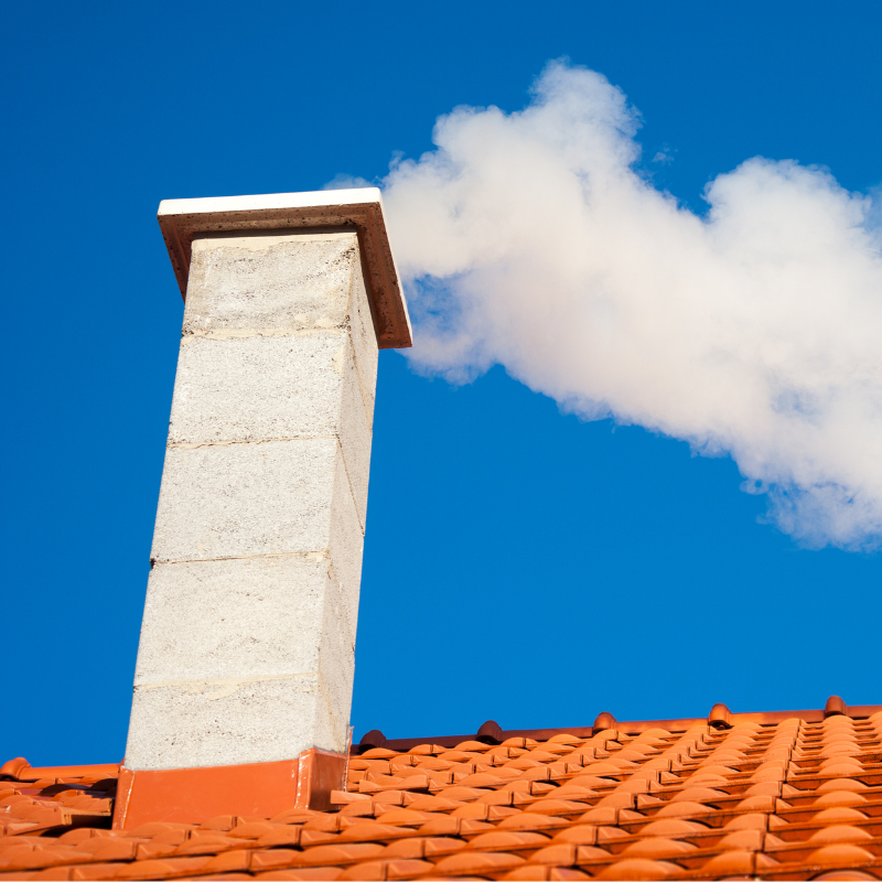 A white chimney with smoke coming out of it sitting on a tile roof with a blue sky in the background.
