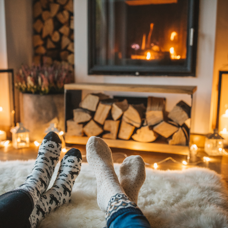 Two sets of feet propped up in front of a wood burning fireplace has wood stacked in front and to the side with little twinkling lights running across the floor.