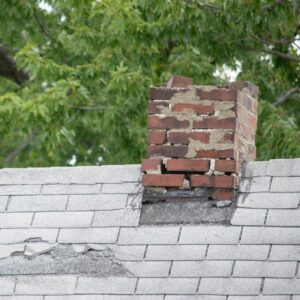 a crumbling masonry chimney on a gray roof with trees in the background