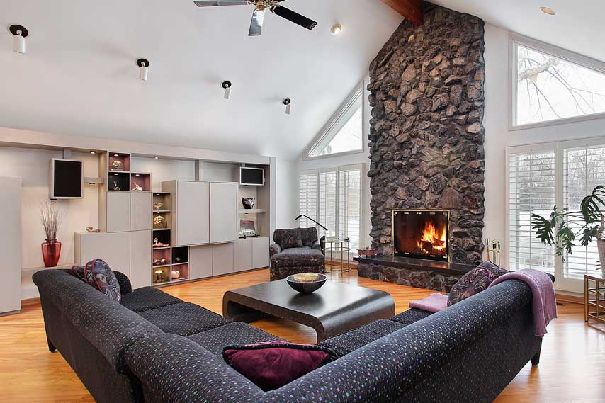 Large stone fireplace in a living room with peaked ceiling and windows on each side, an L shaped sofa, coffee table and chair with cabinets on left wall.