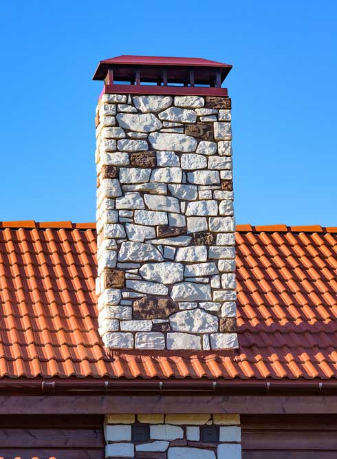 Beautifully restored historic chimney with rock masonry and a chimney cap on a tile roof with a blue sky in the background.